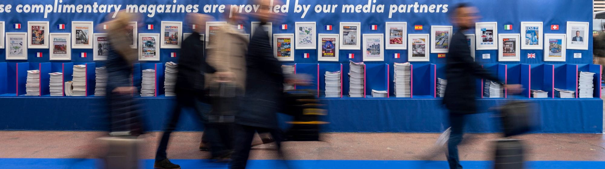 des visiteurs avec des valises traversant un salon commercial et passant devant un mur international de documentation