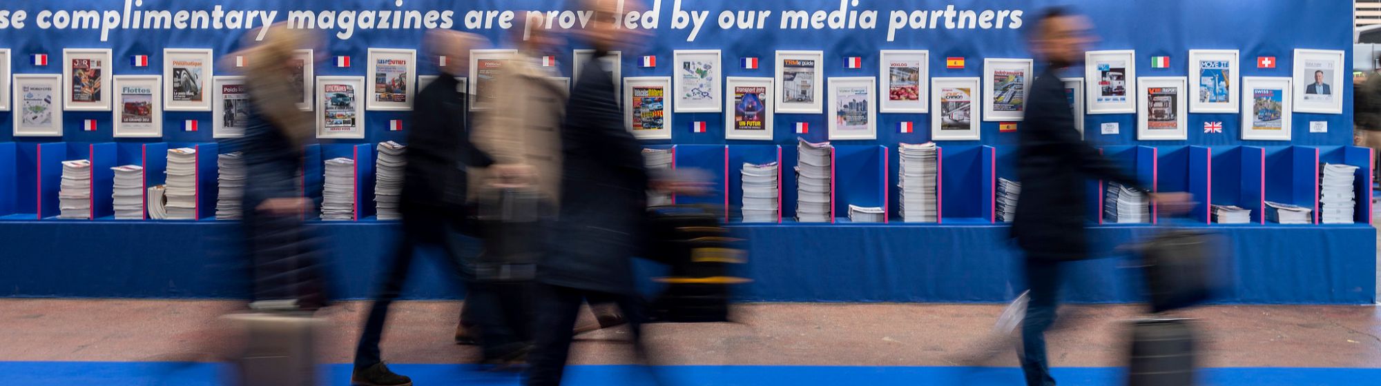 visitors with suitcases walking through a trade fair and past an international wall of documentation